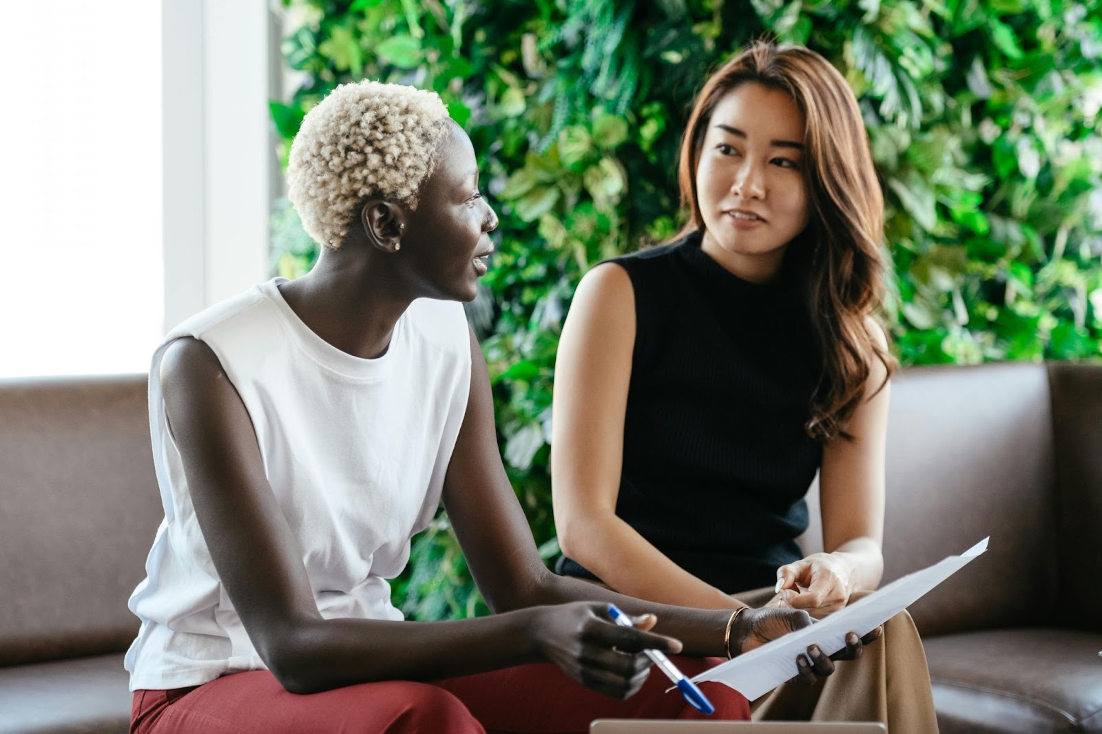 Two people sit side by side on leather couches talking to each other, one of them holding a pen and sheet of paper. 