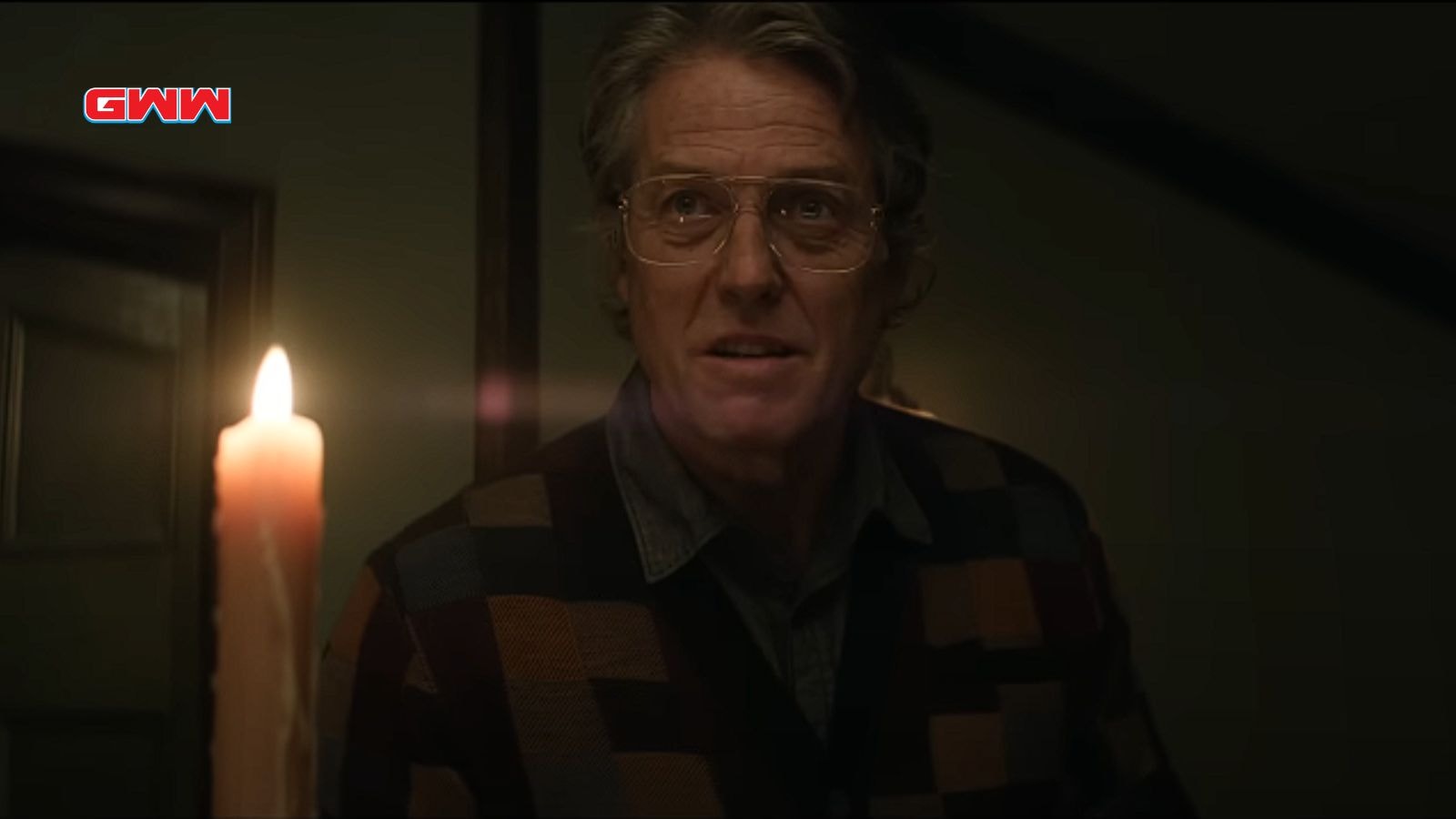 Hugh Grant holding a candle, looking cautious in a dimly lit room.