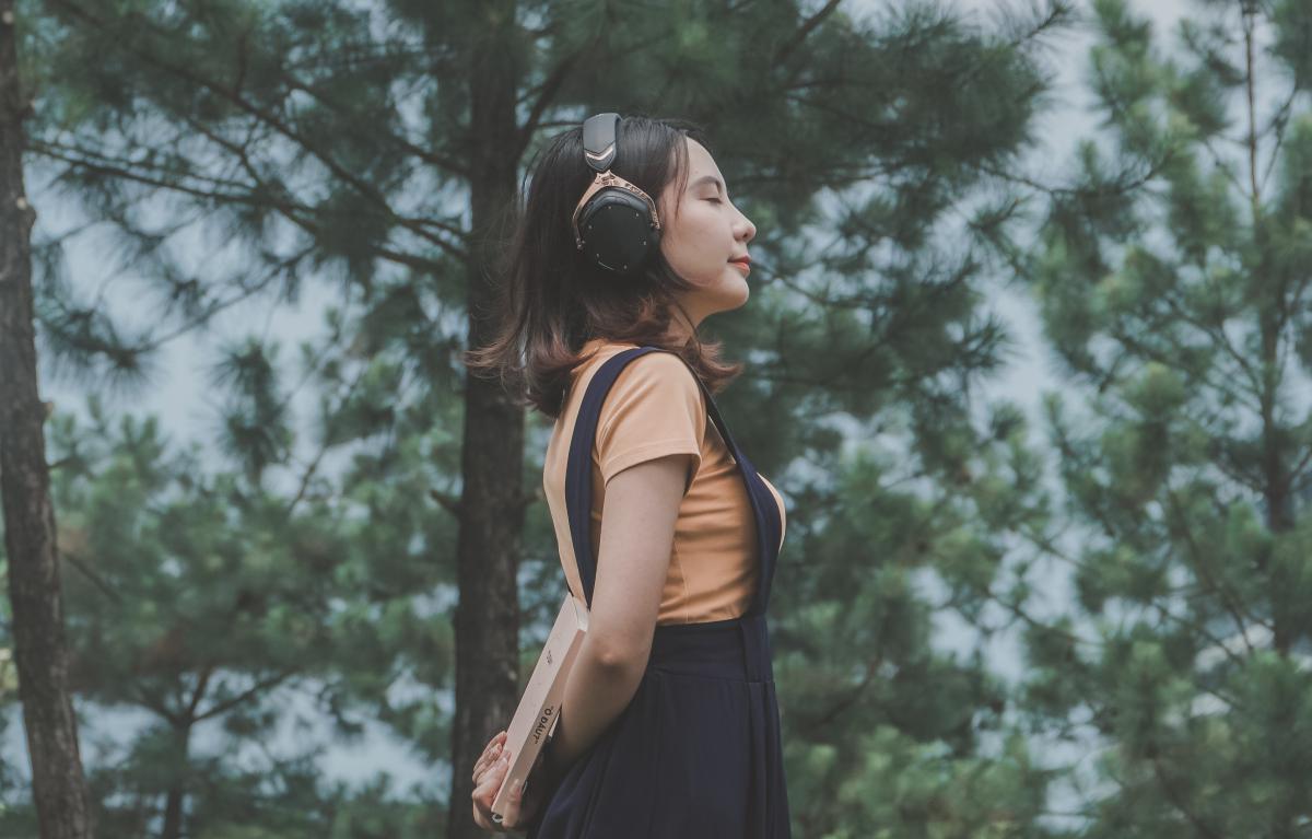 Photograph of a girl wearing heaphones and holding a book behind her with her eyes closed, standing in front of trees