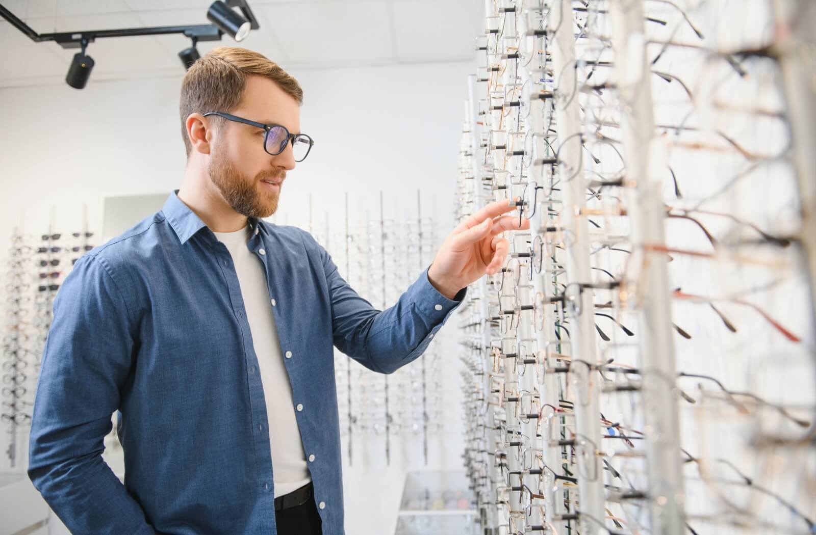 A patient wearing a blue button-down and glasses looks at a wall of different eyeglass styles in a brightly lit clinic to pick a properly fitted frame.