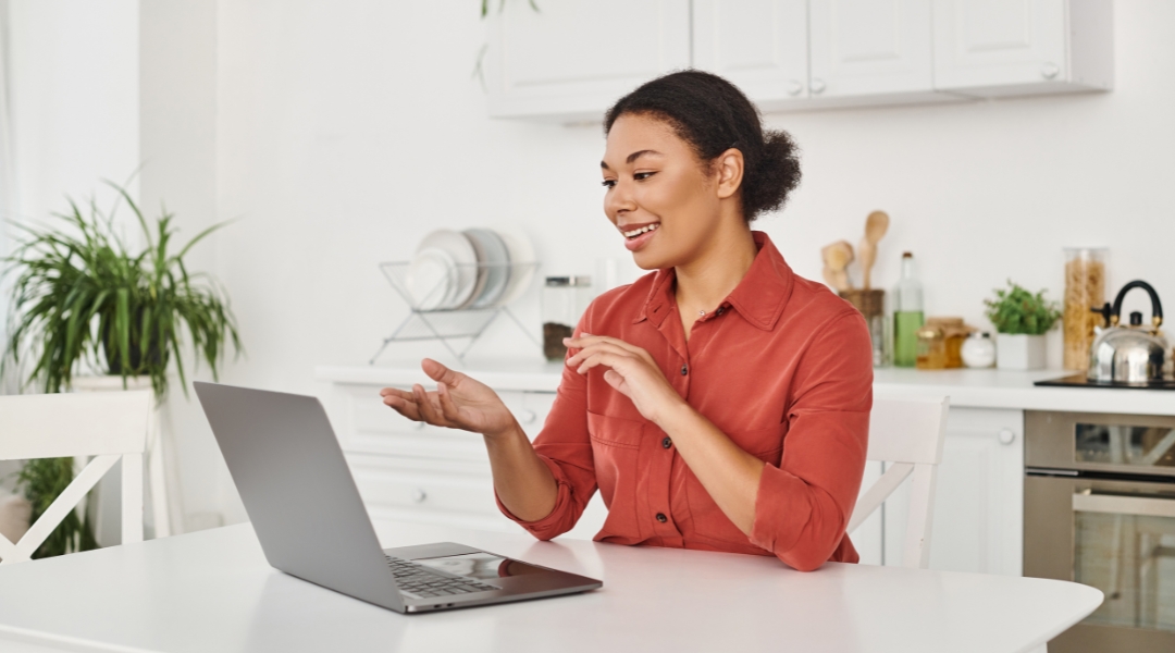 Une nutritionniste diététiste souriante en chemise orange utilise un ordinateur portable dans une cuisine lumineuse pour faire sa consultation en nutrition en visioconférence.