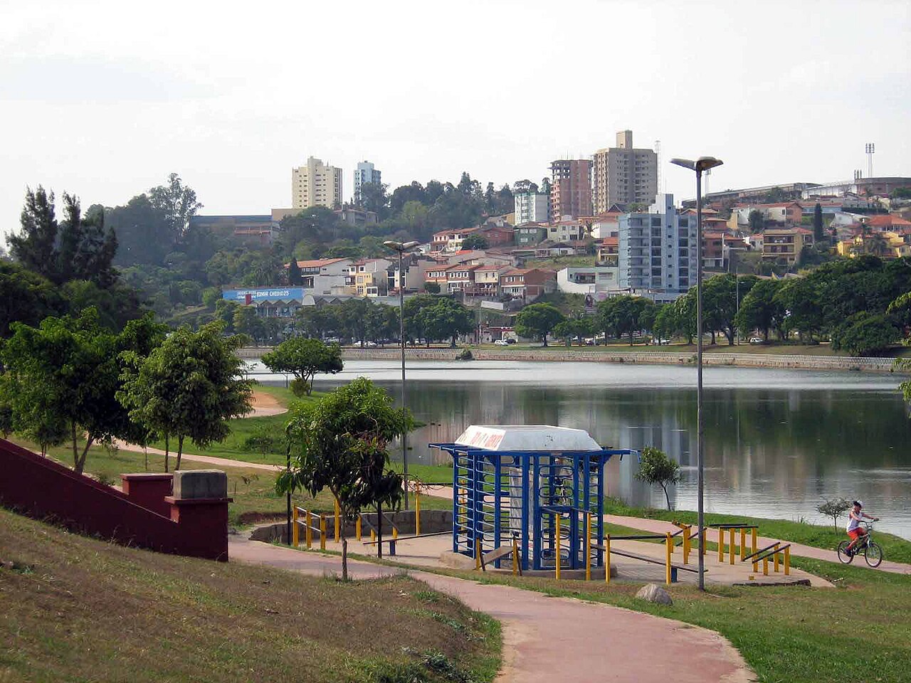 Praça com área de lazer, ciclovia, árvores, lago e cidade ao fundo, em um dia nublado em uma cidade brasileira.