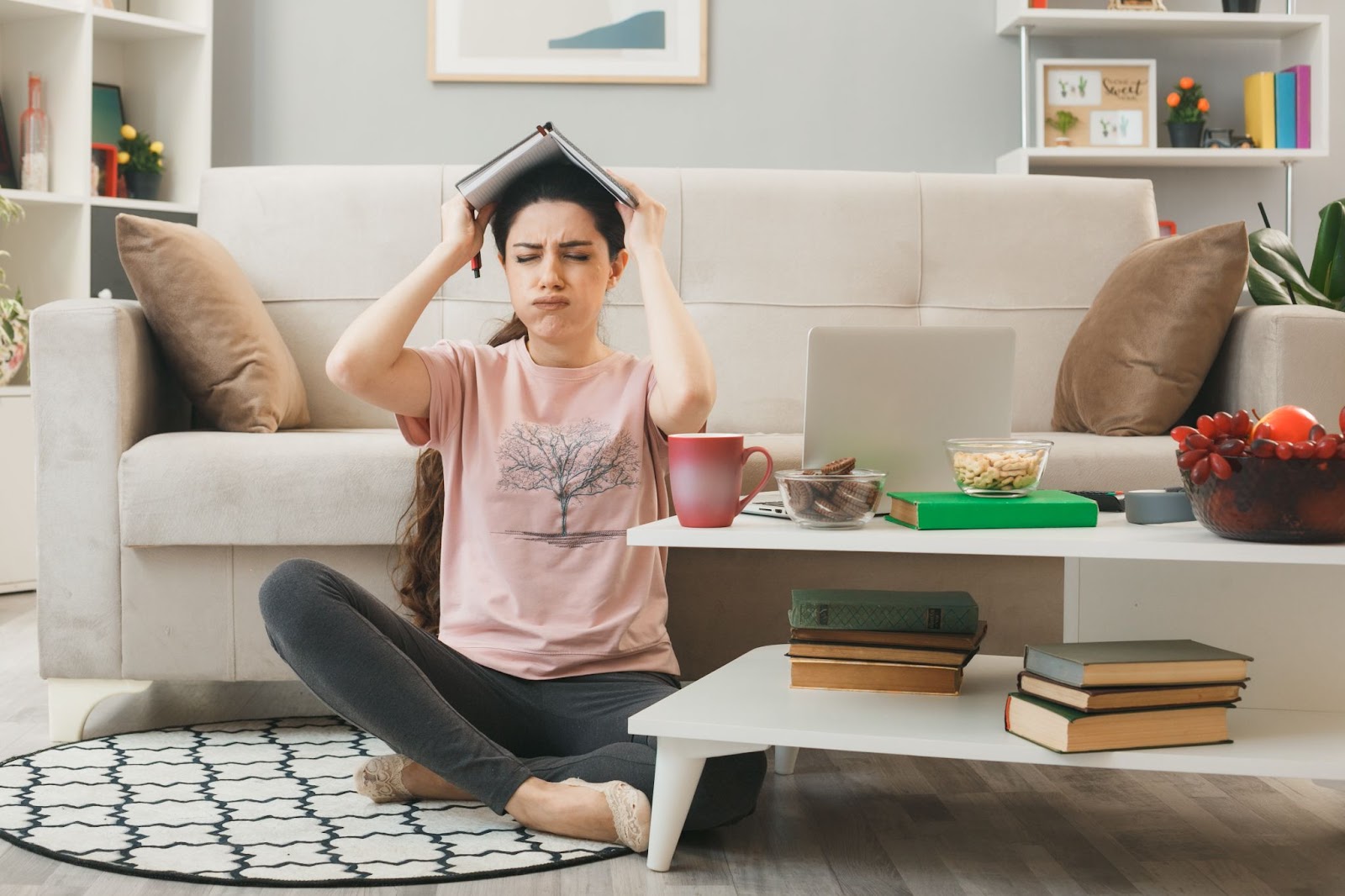 Unpleased with closed eyes, young woman sitting on the floor with book on her head.