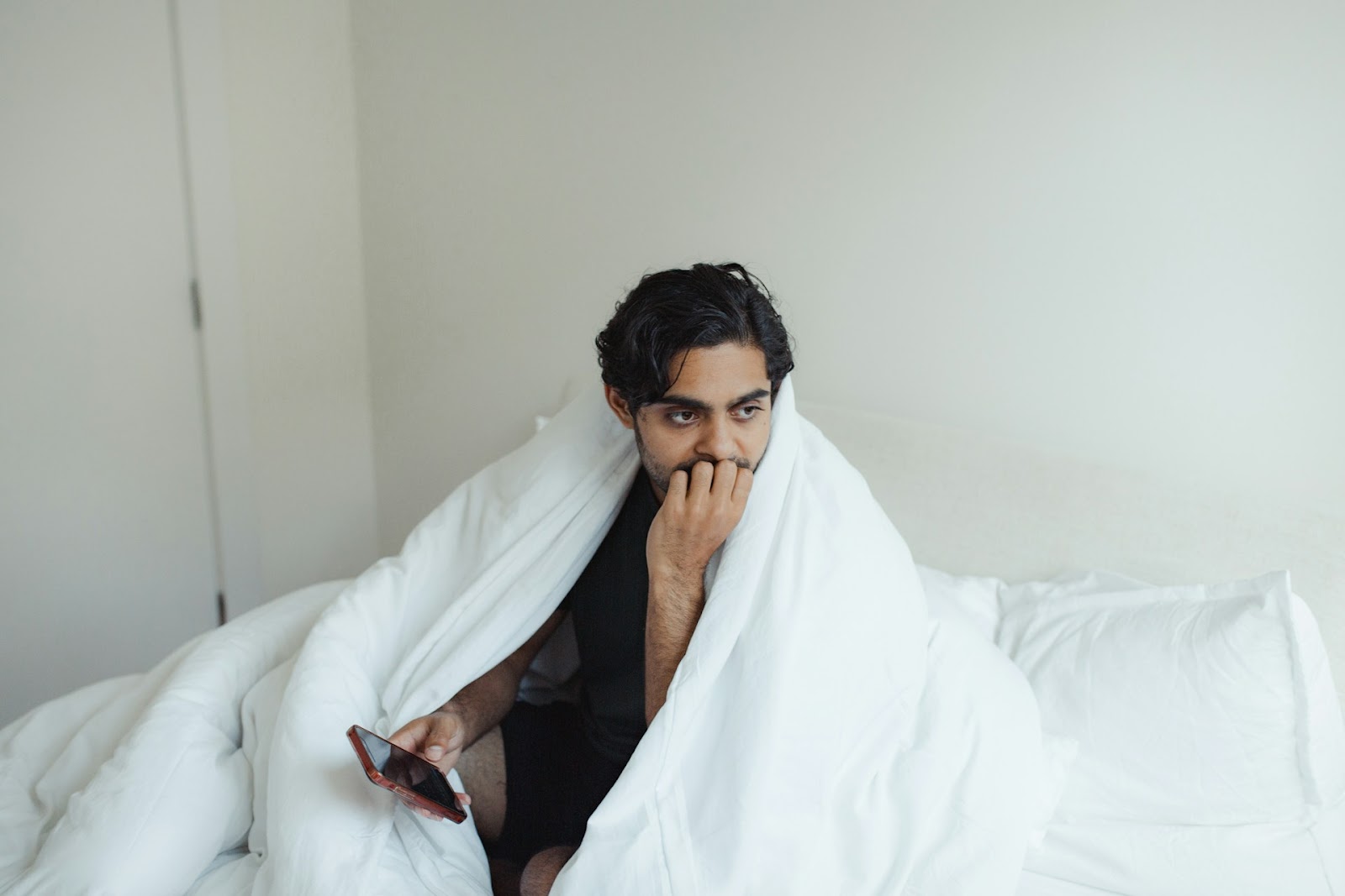 A man sits in bed under his duvet, looking concerned, biting nails, holding his phone in his hand