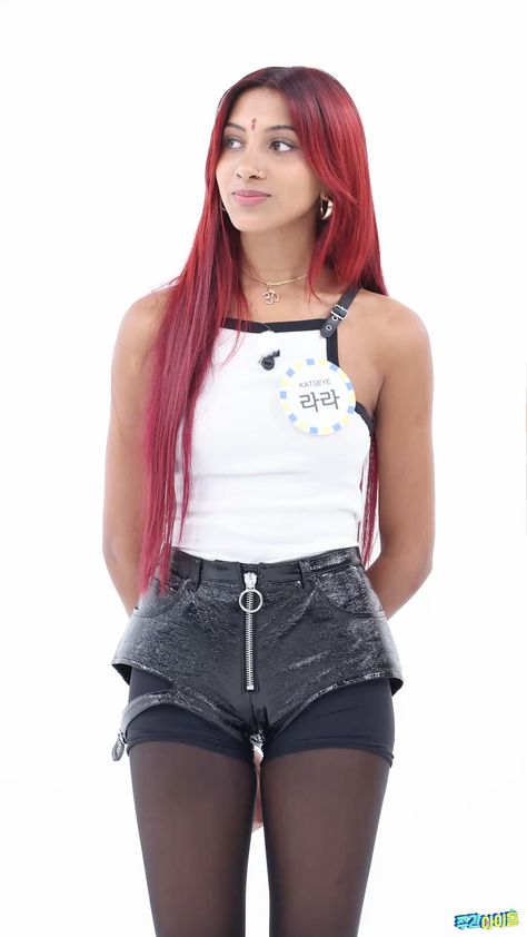 This contains an image of KATSEYE Lara with long red hair wearing black shorts and a white tank top is posing for the camera