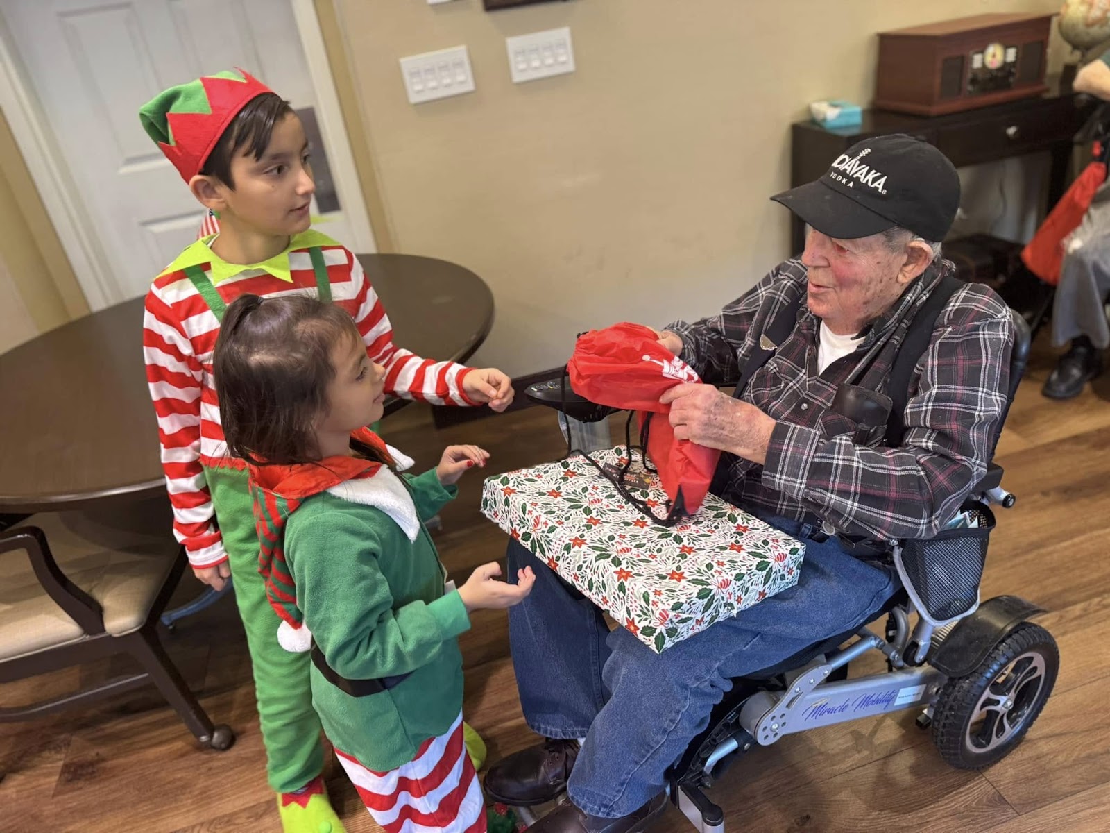 Two children dressed in Christmas clothing receiving a gift from an elderly individual.