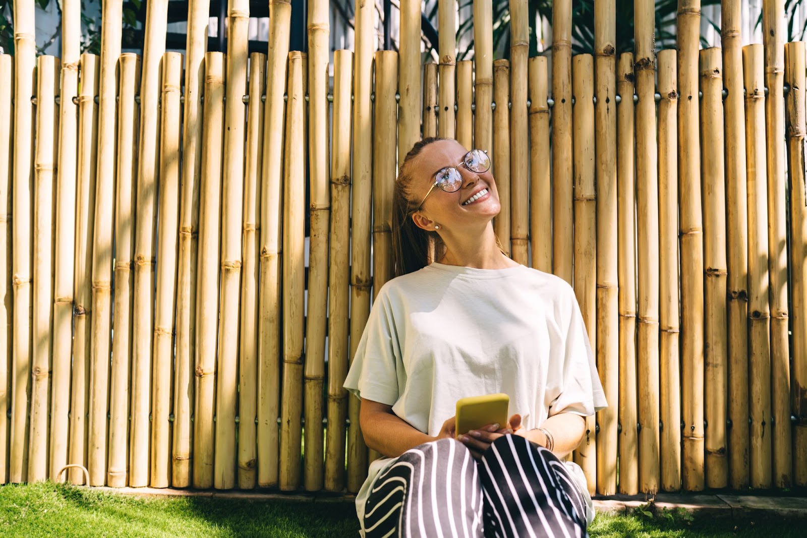 A millennial hipster girl with a smartphone in hand, casually leaning on a bamboo fence in a natural outdoor setting.