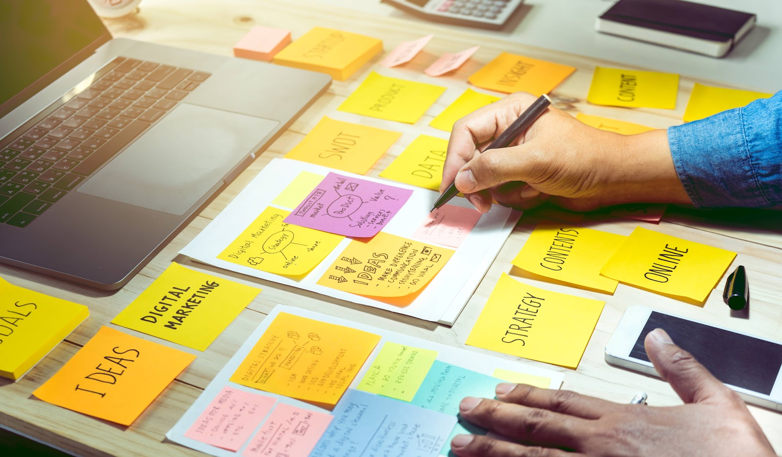 Person writing marketing strategy on sticky notes, laptop nearby on desk