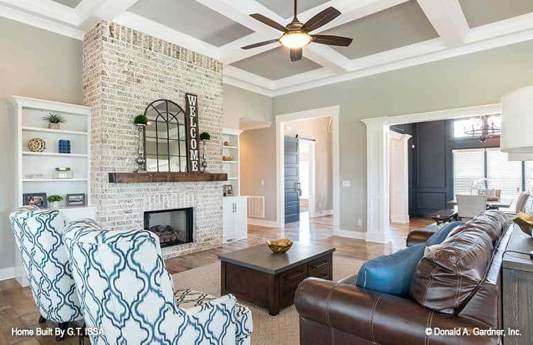 Great room with a coffered ceiling, a leather sofa, patterned armchairs, and a brick fireplace topped with an arched mirror.