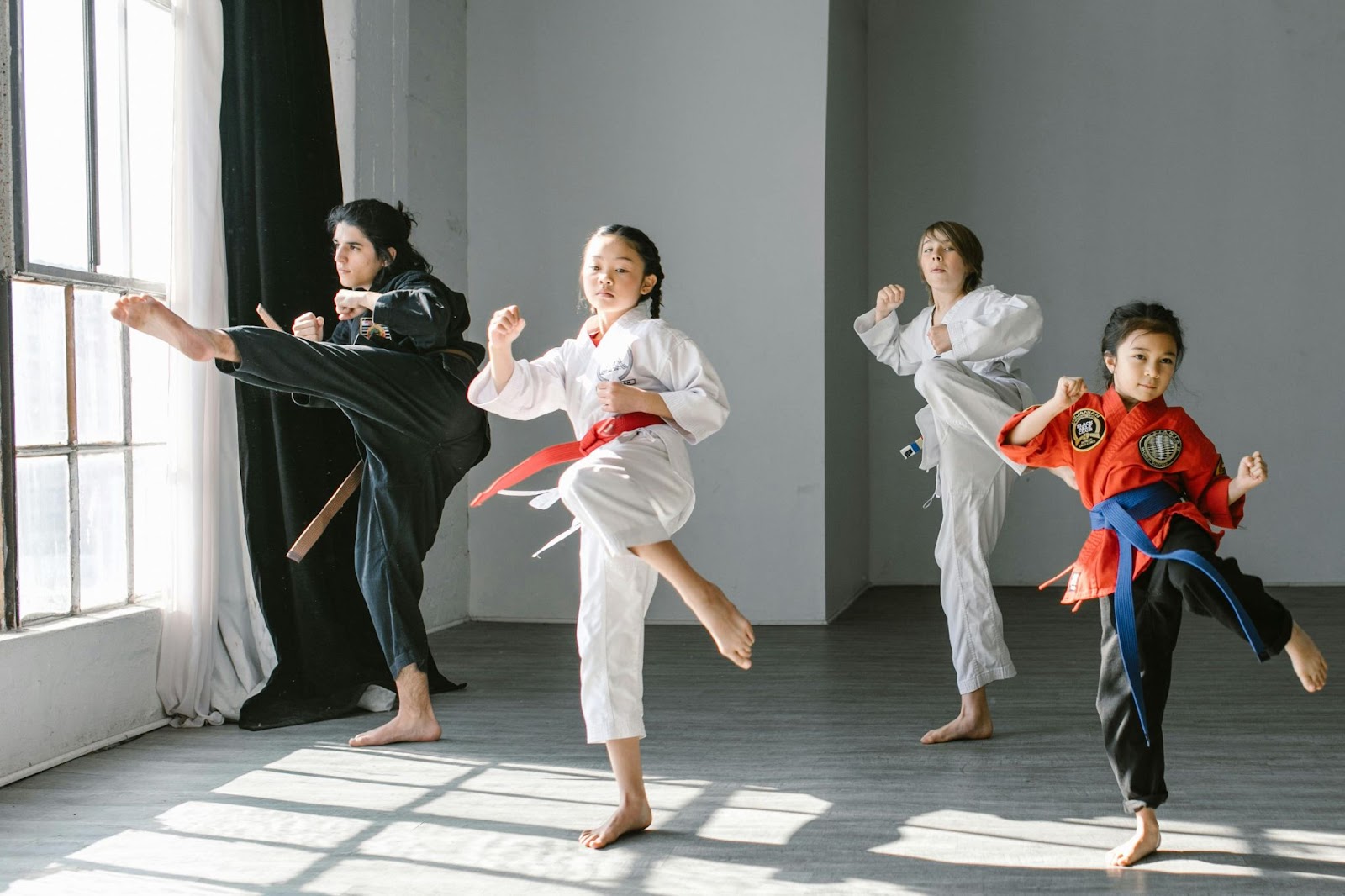 Four young martial arts students performing a synchronized move, each with one leg raised in a balanced stance, wearing traditional uniforms in a bright, open training space.