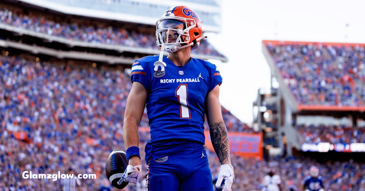 The image shows Ricky Pearsall, a football player for the Florida Gators, standing on the field during a game. He is wearing his blue Gators uniform with the number 1 prominently displayed on his chest and his name above it. Pearsall has a determined expression as he holds a football in one hand. The background is a packed stadium filled with fans, creating an energetic and vibrant atmosphere. The image captures a moment of confidence and focus, with Pearsall looking poised and ready for action.