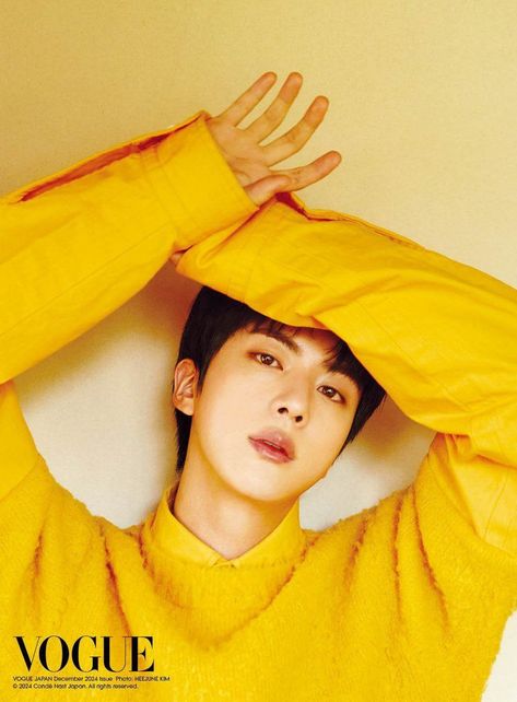 This contains an image of BTS Jin wearing a yellow shirt wiyh his hands on top of his forehead