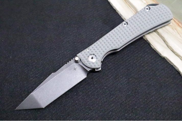 An image showing a Toor folding knife, the Chasm XLT. The knife features a tanto blade and a stonewashed titanium/stealth gray G-10 handle.