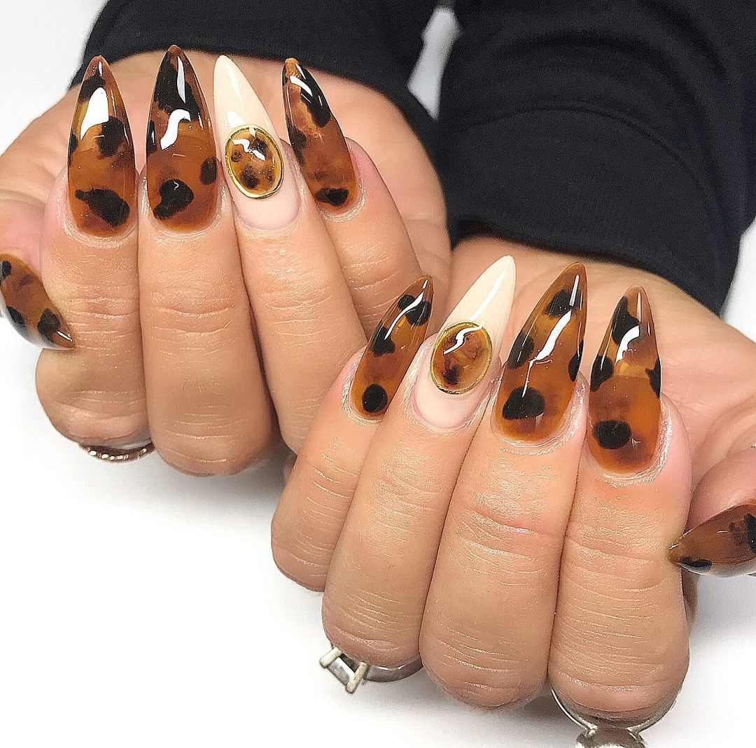 Close-up of a hand with long, almond-shaped nails featuring a glossy brown and white swirl design.
