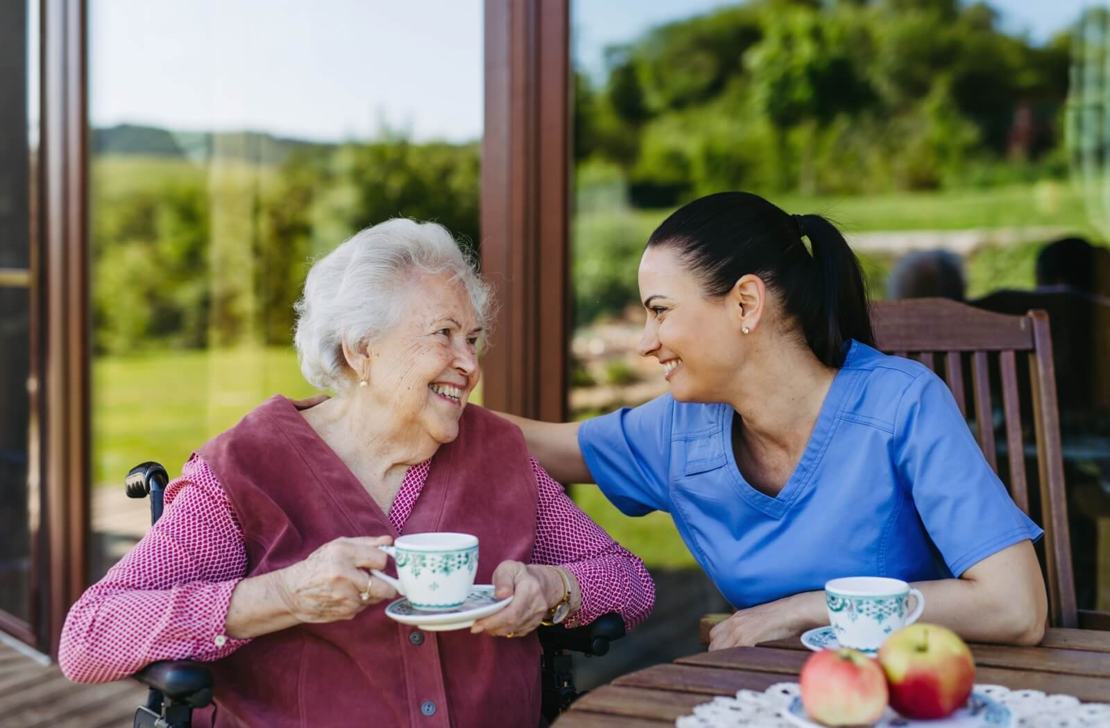 An assisted living resident enjoys a cup of tea on the patio. A caregiver places their arm on the senior's shoulder. Both smile, sharing a joyful moment together.