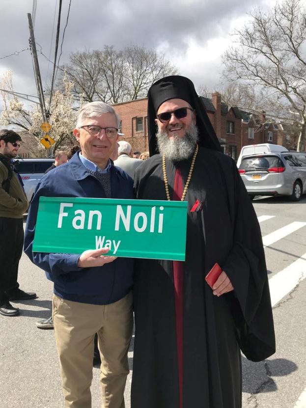 Two men standing on a street holding a sign

Description automatically generated