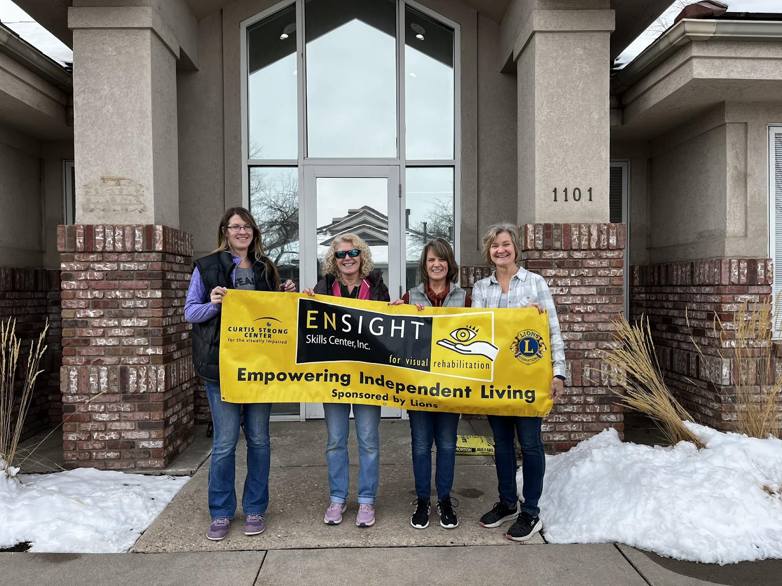 the image shows four women standing outside a building in front of a doorway, holding a yellow banner with the text "ENSIGHT Skills Center, Inc. Empowering Independent Living" and logos for "Curtis Strong Center for the visually impaired" and "Lions International." The group is standing on a concrete path with patches of snow on the ground.