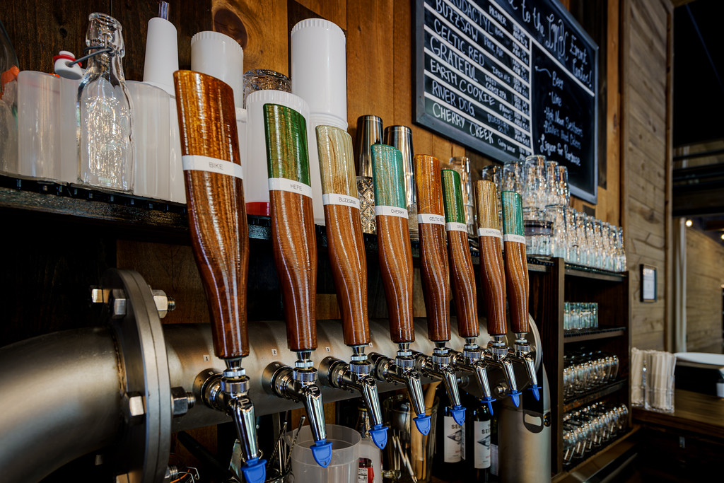 Close-up of a row of wooden beer tap handles attached to a metal tap system. The background features shelves with glassware and a chalkboard menu listing various beer options in Little Miami Brewing company.