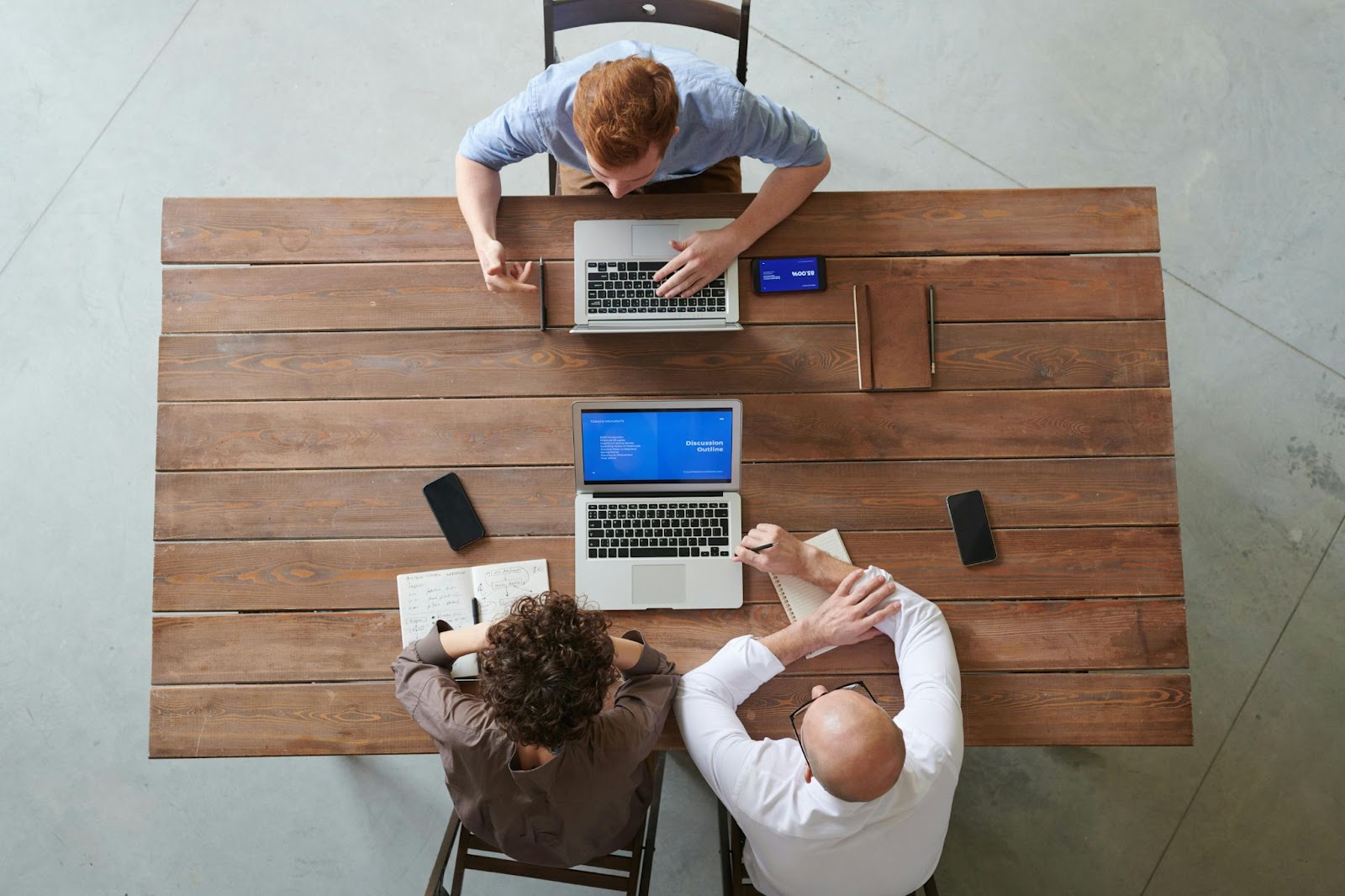 Tres personas trabajando en una mesa de madera con laptops, usando software de gestión, vista desde arriba