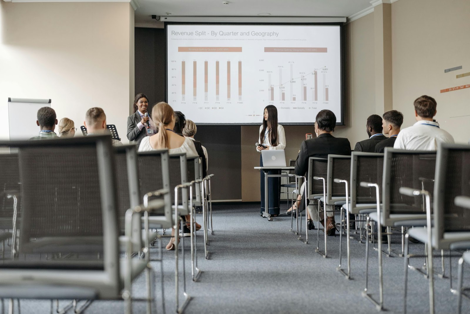 Two women hold a PowerPoint presentation while their coworkers sit at desks.