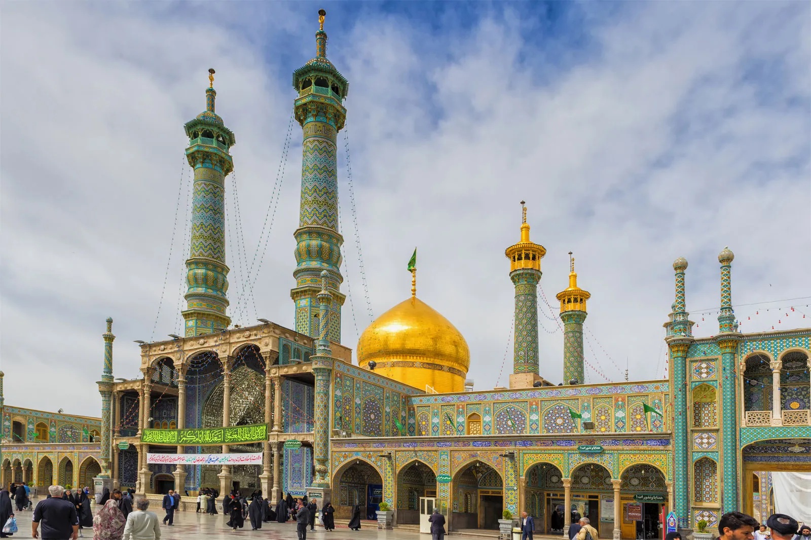 Daytime view of the Shrine of Fatima Masumeh in Qom, Iran, featuring golden domes, intricate turquoise minarets, and beautifully tiled facades with worshippers in the courtyard.