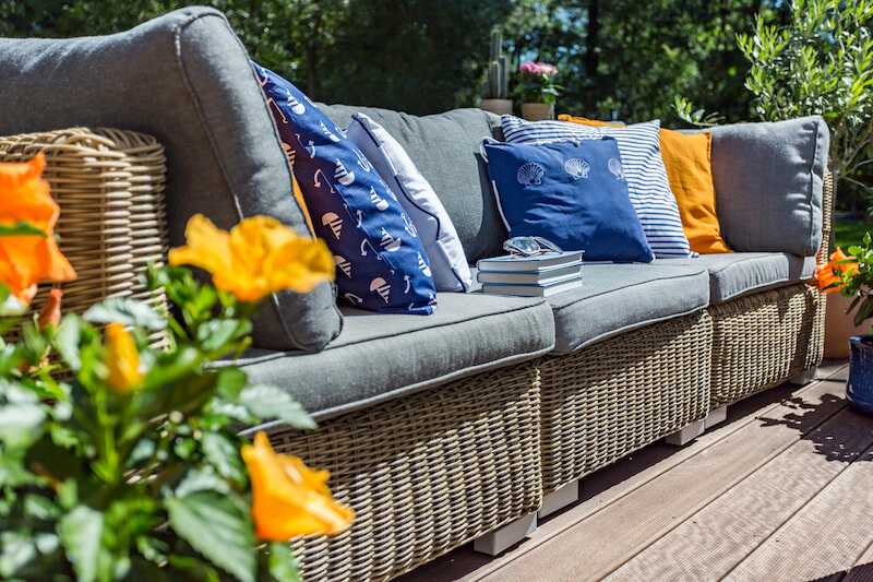 A stylish outdoor wicker sofa is adorned with colorful and patterned cushions, creating a cozy and inviting atmosphere on the patio. Bright yellow flowers and lush greenery surround the seating area, enhancing the relaxing ambiance of this outdoor furniture setup.