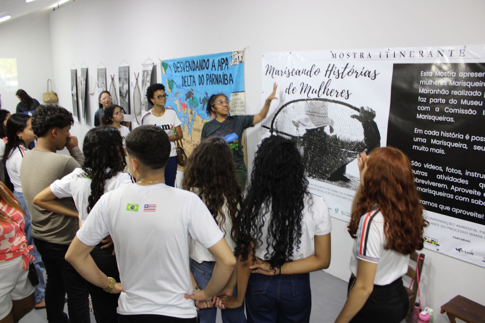 Em uma parede branca, há cartazes e banners da exposição 
"Mariscando Histórias e Mulheres", sobre marisqueiras. Uma mulher está apontando a mão para um dos cartazes, em frente a estudantes.