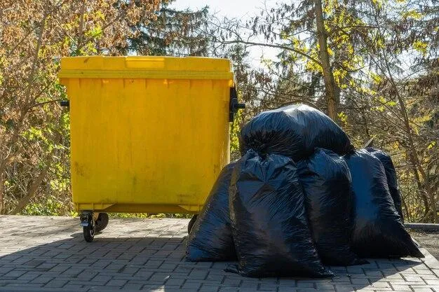 A skip bin sits next to a pile of black garbage bags on a paved surface. The skip bin has wheels for mobility, while several filled black plastic garbage bags are stacked beside it awaiting collection.
