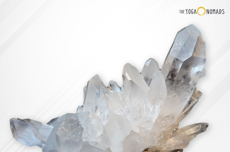 A cluster of clear quartz crystals with pointed terminations and varying degrees of transparency. The crystals are positioned against a light background that fades from white to beige, with a diagonal stripe pattern.