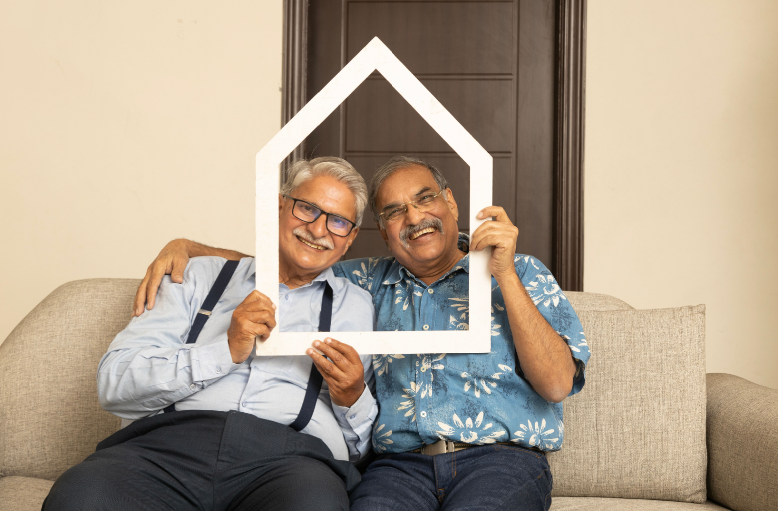 2 senior friends sitting on a cozy couch, holding a white house-shaped frame in front of their faces, smiling.