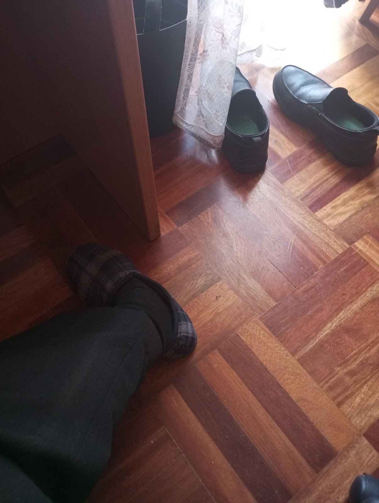 A man's empty black flats on a wooden floor next to white lacy curtains and a wooden door. 