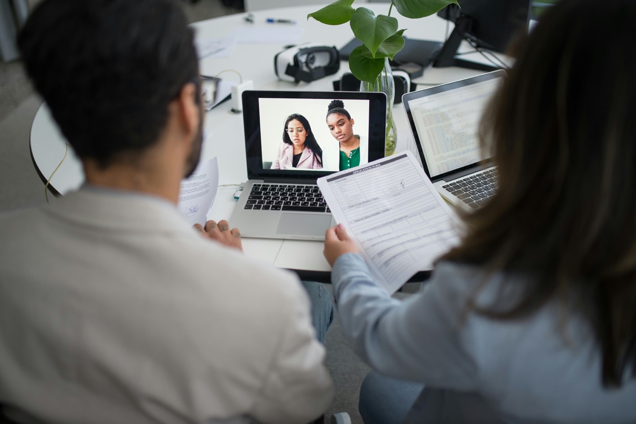 Two professionals conducting a video interview with candidates on a laptop screen while looking at papers.