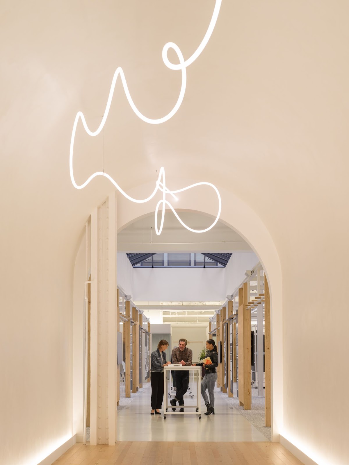 A man and two women stand at a rolling desk in a curved hallway with swirling decorative lighting and light peach-colored walls.