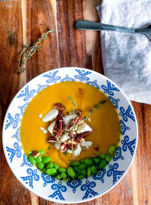 Creamy butternut squash soup garnished with pecans, goat cheese, and edamame beans in a patterned bowl.