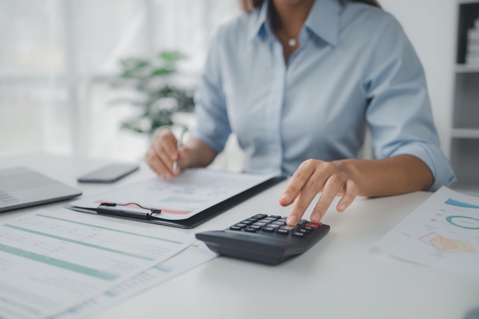 A woman sitting at a desk and using a calculator and a clipboard with various charts on papers to budget.