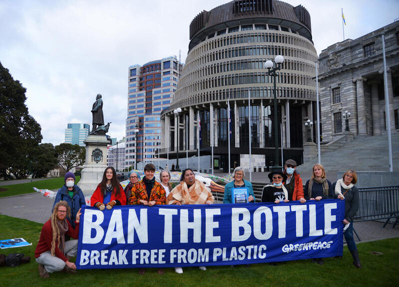 12 people at NZ Parliament hold a big banner that says BAN THE BOTTLE, break free from plastic, Greenpeace.