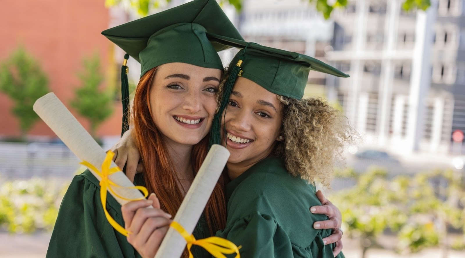 Two smiling nutrition graduates in green caps and gowns hug while holding diplomas outdoors after their ceremony.