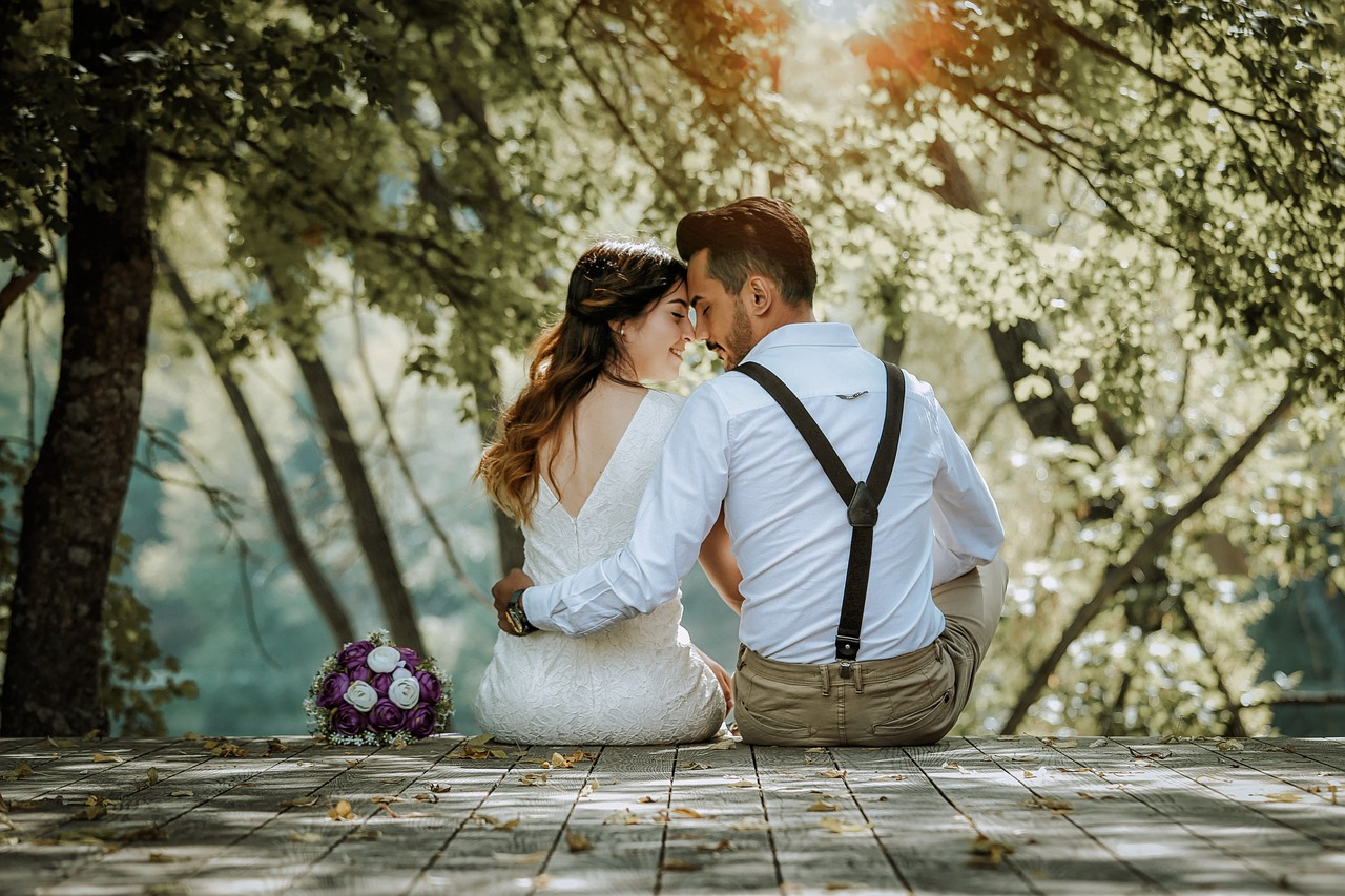 Um casal, com um homem e uma mulher com vestimentas relacionadas a casamento, estão de costas se abraçando de lado, com os rostos encostando