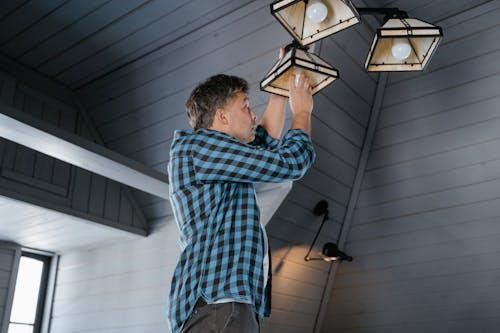 Free A man in a plaid shirt changes a light bulb on a ceiling lamp in a stylish, modern interior. Stock Photo