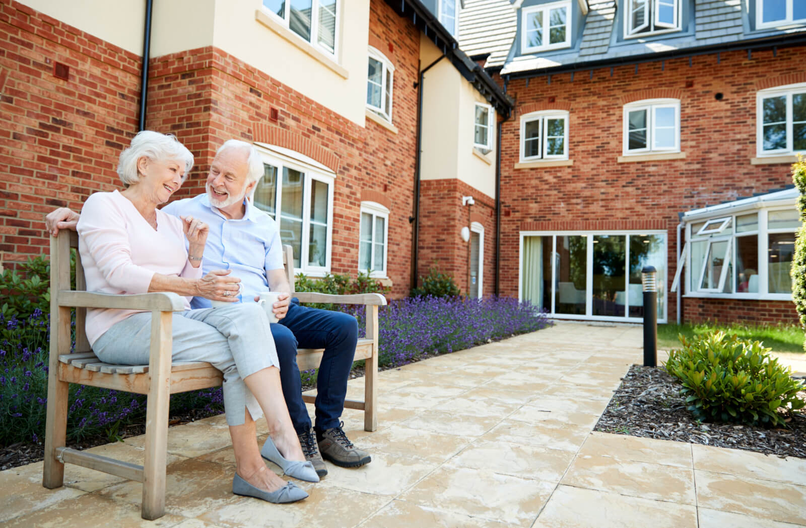 An older married couple sitting on a wooden bench in the common area outside a senior living community.