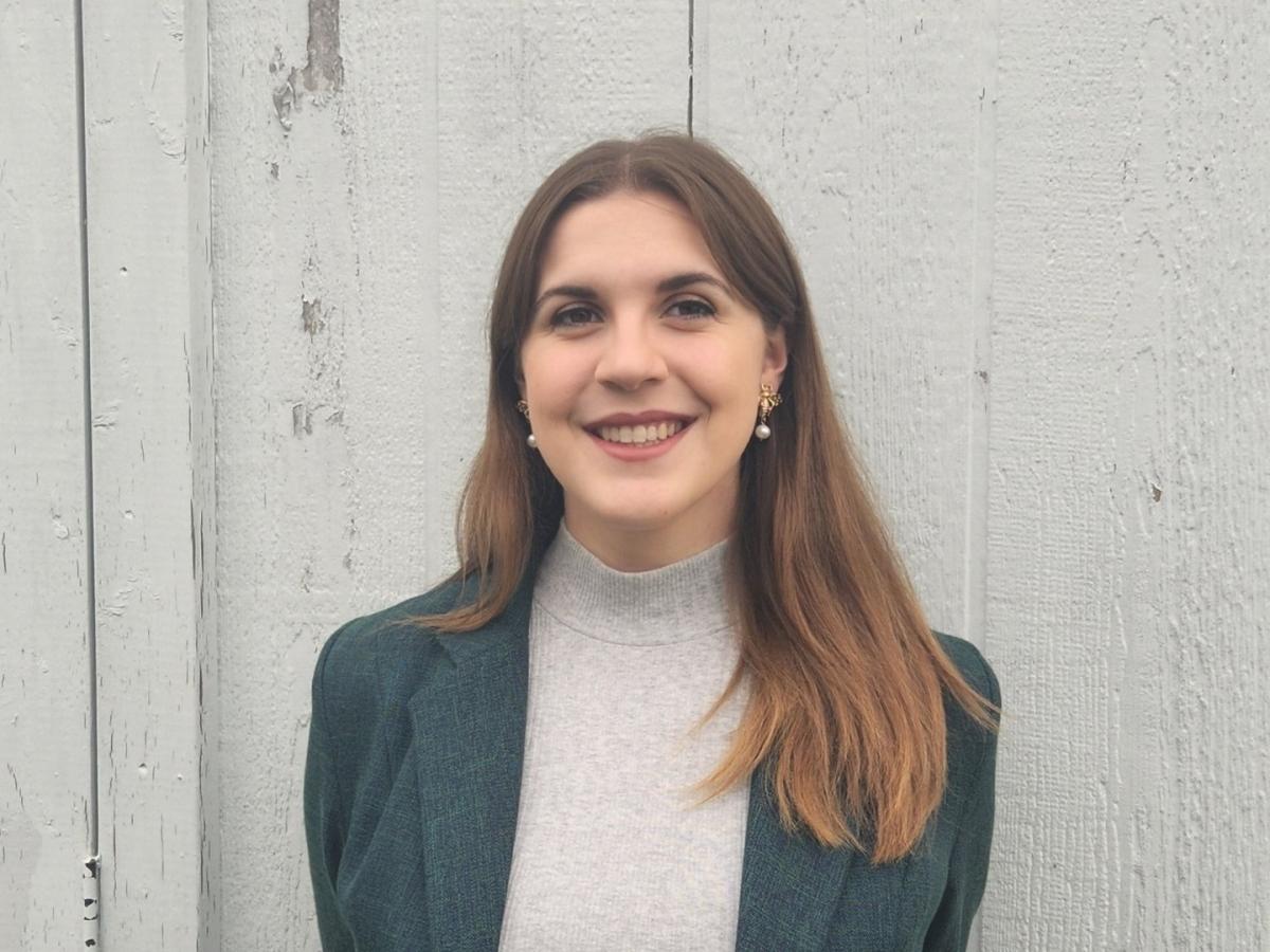 Andrea Ens standing in a grey sweater and dark blue jacket, smiling at the camera.