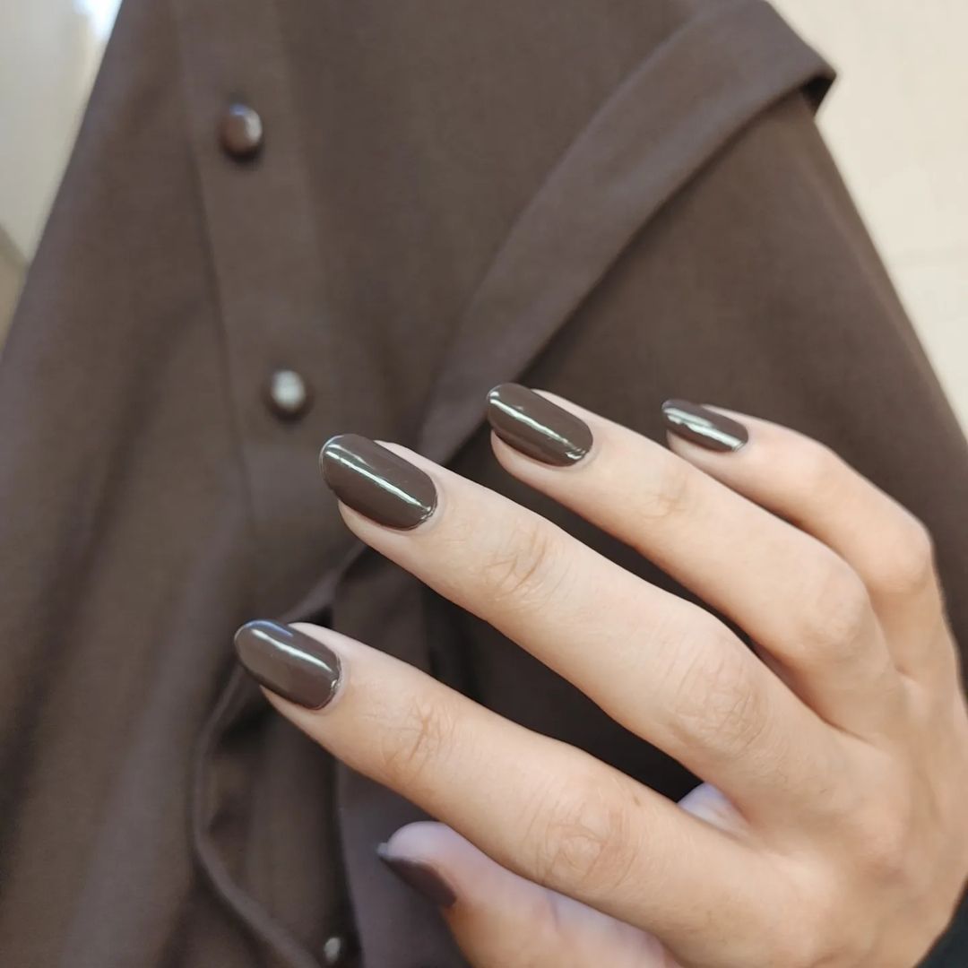 Close-up of a hand with brown polished nails against a brown coat with visible buttons.

