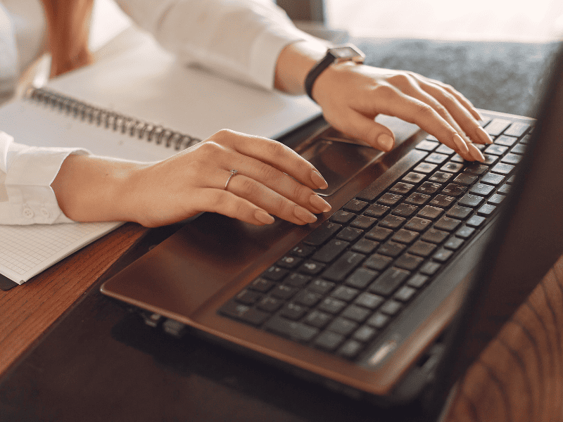 Pessoa digitando em um notebook com caderno de anotações ao lado.

