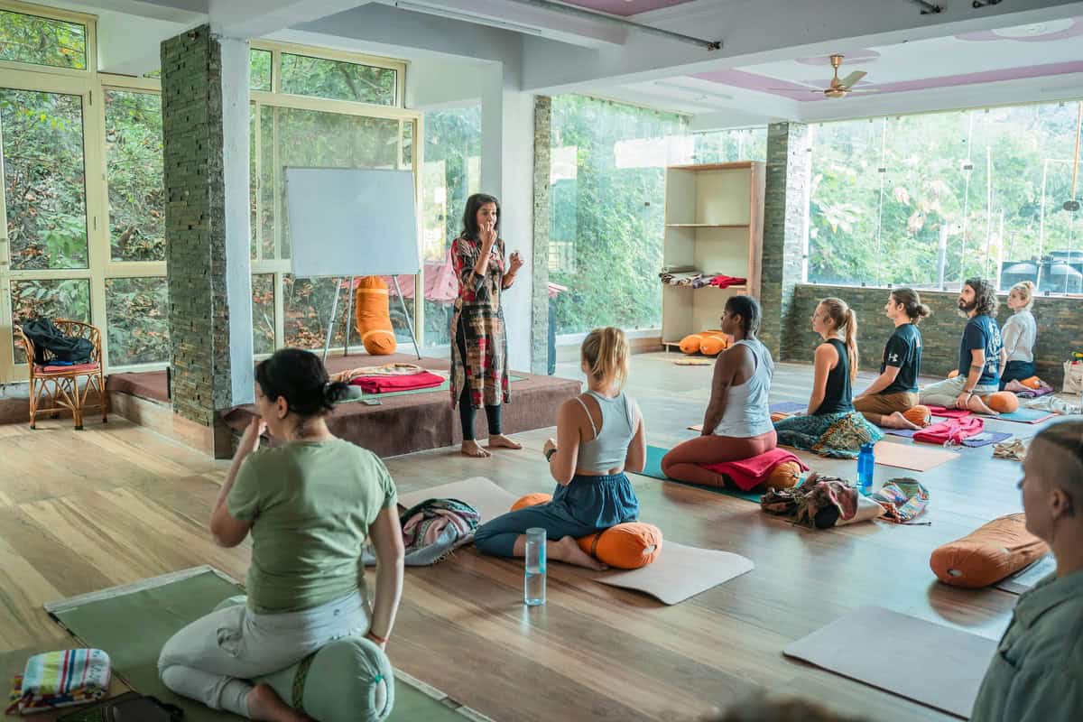 Foto de um estúdio de yoga com várias pessoas fazendo yoga sentadas em cima de Bolster de yoga que é uma almofada cilíndrica macia que facilita o exercício.