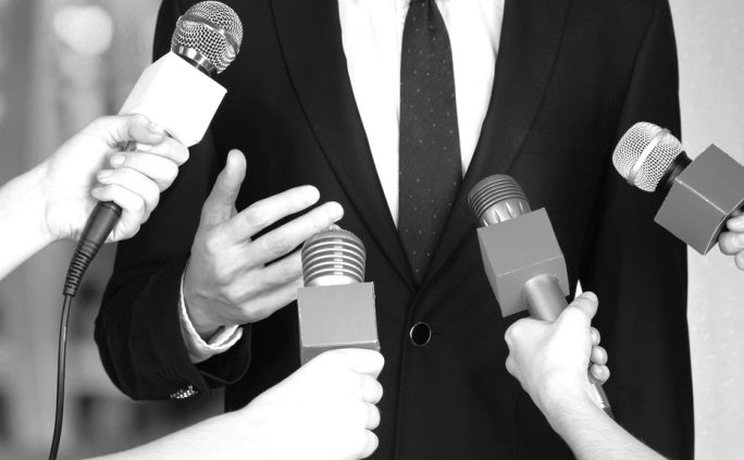 A person in a suit being interviewed by several reporters holding microphones.