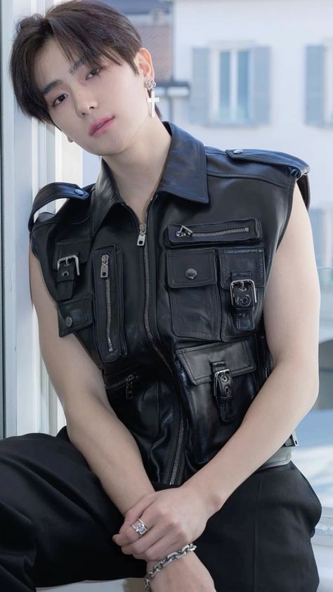 This contain an image of  THE BOYZ's Hyunjae, wearing a black leather vest sitting on a window sill with his hands on his leg