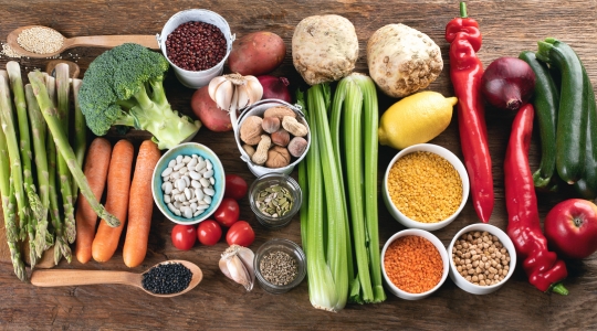 An assortment of fresh vegetables and seeds, including asparagus, broccoli, carrots, celery, red peppers, zucchini, tomatoes, garlic, potatoes, celeriac, lemon, nuts, and various seeds and legumes, arranged on a wooden table.