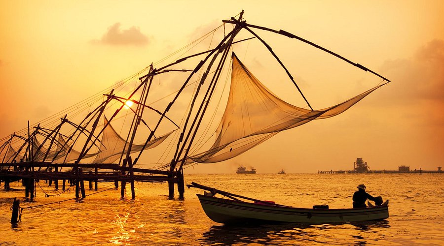 Traditional Chinese fishing nets silhouetted against a golden-orange sunset over the waters of Fort Kochi, Kerala. A lone fisherman sits calmly in a wooden boat, adding a serene human element to the tranquil coastal scene, with ships visible on the distant horizon.

