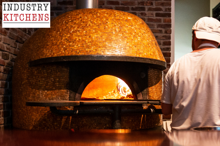 Professional brick pizza oven with golden flames inside and cook preparing to place food, ideal for wood-fired cooking