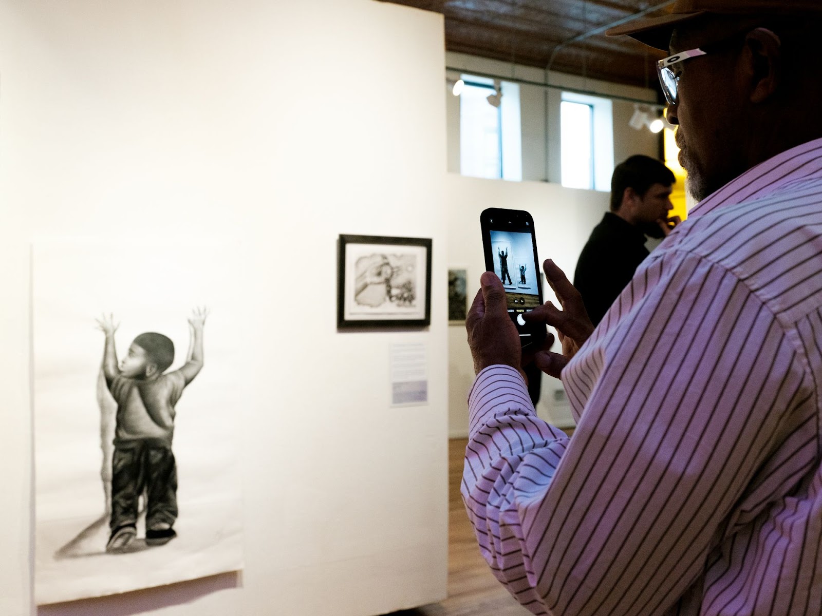 Image: Suspects by Mike Sullivan installed at exhibition More Beautiful, More Terrible: Human of Life Row. A Black person takes a photo of an illustration of a Black child standing with their hands up against the wall, but looking to the left. Photo by Olivia Mikolai Ridge, 2024.